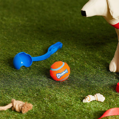 Compact blue Chuckit! ball launcher and orange tennis ball resting on grass beside a dog display figure and other dog toys.