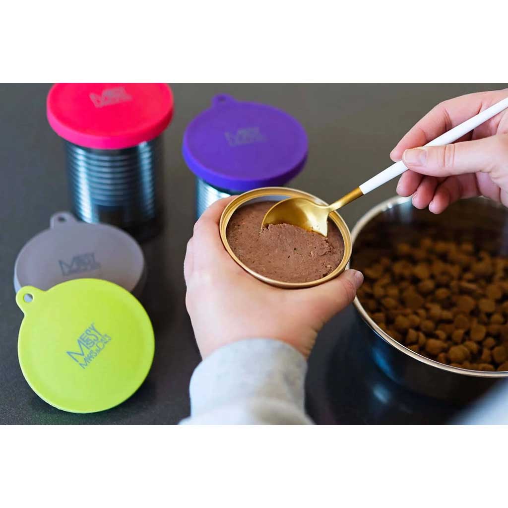 Wet pet food being spooned from a can beside colorful Messy Mutts silicone can covers and a bowl of kibble.