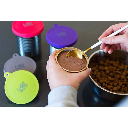 Wet pet food being spooned from a can beside colorful Messy Mutts silicone can covers and a bowl of kibble.