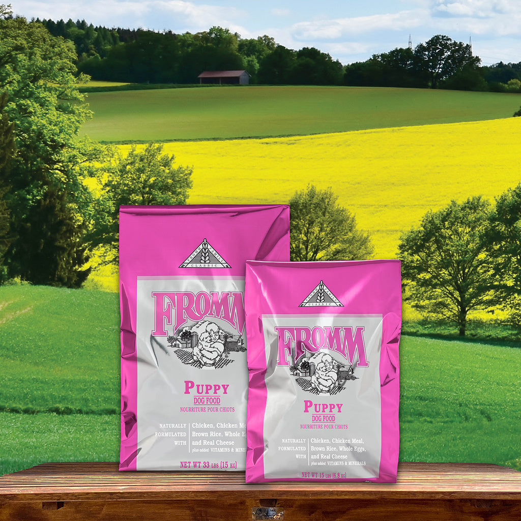 Two pink and silver Fromm Classic Puppy dry dog food bags on a wooden ledge overlooking green fields
