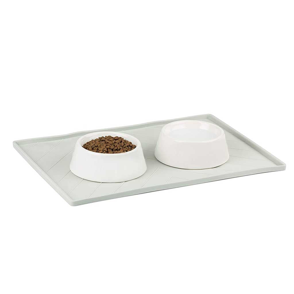 Two white pet bowls, one filled with kibble, resting on a gray Messy Mutts silicone food mat.